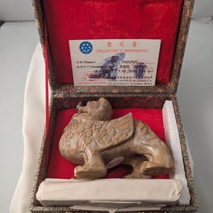 May include: A small, intricately carved stone sculpture of a mythical creature with wings, in a wooden box with a red velvet lining. The box has a label that reads "Collector's Certificate" and includes information about the geological age of the fossil.  The sculpture is a scientific souvenir from the Nanjing Institute of Geology & Paleontology, Academia Sinica.