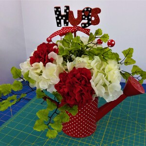 May include: A red watering can with white polka dots is filled with white and red flowers. The flowers are surrounded by green leafy branches. A wooden sign with the word "Hugs" in black and white polka dots is placed on top of the arrangement.