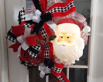 May include: A festive Christmas wreath featuring a light-up Santa Claus face and an assortment of red, black, white, and checkered ribbons. The wreath is designed for holiday decor and is displayed on a white door.