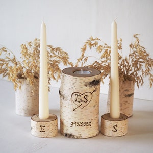 May include: A set of three birch wood candle holders with a heart and initials "E&S" and the date "9/10/2022" engraved. The center holder has a small tea light candle. The two outer holders have tall white candles.