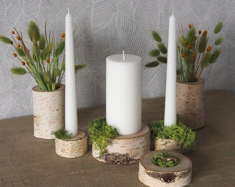 May include: A collection of rustic birch wood candle holders and vases. The arrangement includes white candles of varying sizes, green moss, and decorative grass. One holder displays a pair of rings.