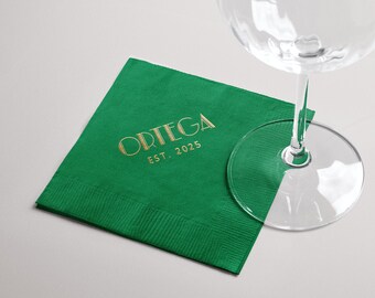 Pode incluir: Uma guardanapo de papel quadrado verde esmeralda com letras douradas que dizem "ORTEGA EST. 2025". Um copo de vinho transparente é parcialmente visível no fundo. O guardanapo é dobrado e tem uma borda texturizada.