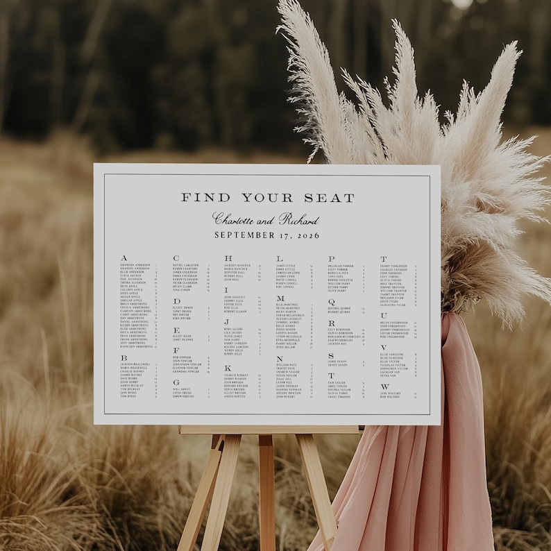 May include: A white seating chart on a wooden easel with the text "Find Your Seat" and the names "Charlotte and Richard" and the date "September 17, 2026". The chart is surrounded by tall, dried grasses and a pink fabric.