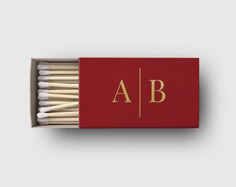 May include: A red matchbox with gold lettering and a vertical line separating the letters "A" and "B". The matchbox is open, revealing a row of wooden matches with white tips. The matches are arranged neatly inside the box.