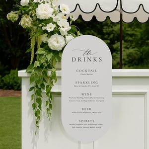May include: An oval-shaped white sign with black text reading The Drinks, Cocktail, Sparkling, Wine, Beer, and Spirits, listing various beverage brands. The sign is attached to a white bar counter adorned with a large arrangement of white flowers.
