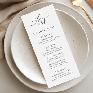 Può includere: Un menu di nozze rettangolare bianco con testo nero in carattere serif, che presenta il monogramma AW, la data 18 ottobre 2027 e un elenco di portate tra cui insalata Caesar, filetto di manzo, petto di pollo, branzino e torta nuziale.