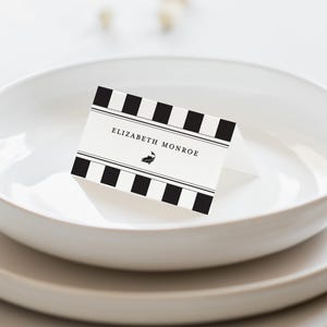 May include: A folded place card with the name "ELIZABETH MONROE" printed in black on a white background. The card features a black and white striped pattern at the top and bottom. The card is placed on a white plate.