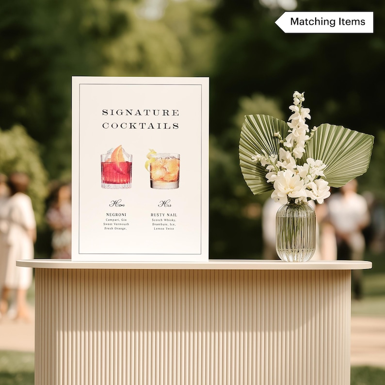 May include: A white framed sign with "Signature Cocktails" text, featuring watercolor illustrations of two cocktails. A vase of white flowers and greenery sits on a light-colored bar. The sign details a Negroni and a Rusty Nail.
