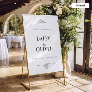 May include: A white wedding welcome sign with gold metal frame. The sign reads "Welcome to the Wedding of Daisy & Oliver" with the date and location. The sign is decorated with art deco style lines and shapes.