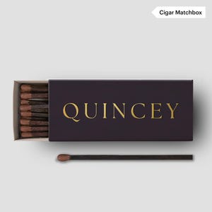 May include: A partially open box of matches with the word "QUINCEY" in gold lettering. The matchbox is dark purple, and the matches have brown tips. The text "Cigar Matchbox" is in the upper right corner.