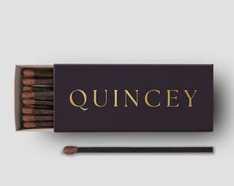 May include: A partially open box of matches with the word "QUINCEY" in gold lettering. The matchbox is dark purple, and the matches have brown tips. The text "Cigar Matchbox" is in the upper right corner.