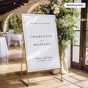May include: A gold metal wedding sign with a white background and black text. The sign reads "WELCOME TO OUR WEDDING", "CHARLOTTE AND RICHARD", and "Charleston, South Carolina, September 17, 2016". The sign is surrounded by greenery.