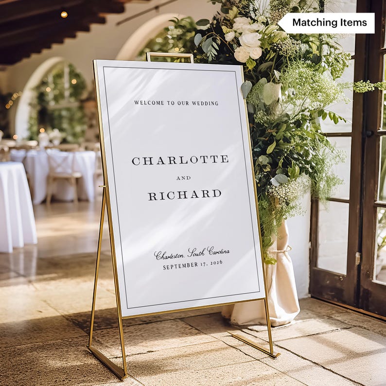 May include: A gold metal wedding sign with a white background and black text. The sign reads "WELCOME TO OUR WEDDING", "CHARLOTTE AND RICHARD", and "Charleston, South Carolina, September 17, 2016". The sign is surrounded by greenery.