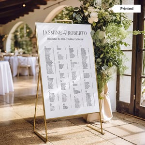 May include: A white seating chart with gold metal frame displays names for a wedding. The text at the top reads "Jasmine & Roberto" with the date December 31, 2026, and location Malibu, California. The chart is surrounded by floral arrangements.
