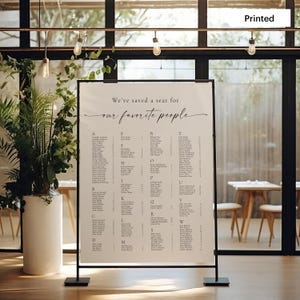 May include: A white seating chart with a black frame, printed with the text "We've saved a seat for our favorite people". The chart is displayed on a black stand in a modern setting.