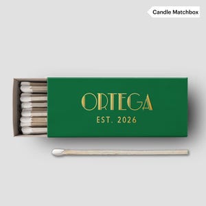 May include: A green matchbox with the word "ORTEGA" in gold lettering and the text "EST. 2026". The matchbox is open, revealing several matches with white tips. One match is lying outside the box.