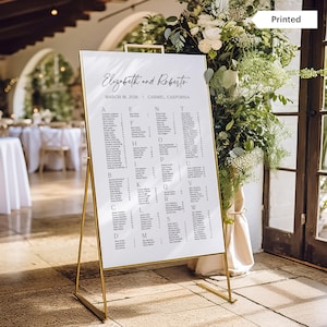 May include: A white wedding seating chart with a gold frame on a gold easel stand. The chart lists guest names in alphabetical order by last name. The chart is titled "Elizabeth and Roberto" and includes the date "March 18, 2026" and location "Carmel California".