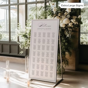 May include: A white wedding seating chart with black text. The chart is on a silver easel and has the words "Welcome Genevieve & Michael" at the top. The chart lists tables and guest names.