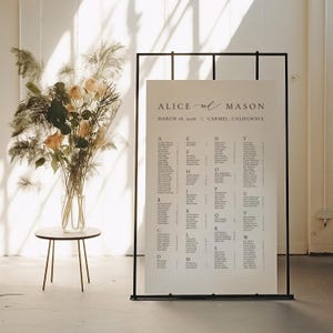 May include: A wedding seating chart with the names of the guests, displayed on a large white sign. The sign is held by a black metal frame. The top of the sign reads "Alice and Mason" and the date and location of the wedding. A vase of flowers sits on a small table.