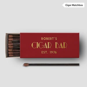 May include: A red matchbox with gold lettering that reads "ROBERT'S CIGAR BAR EST. 1976." The matchbox is open, revealing a row of matches with brown tips. One match is separate from the box.