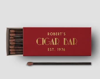 May include: A red matchbox with gold lettering that reads "ROBERT'S CIGAR BAR EST. 1976." The matchbox is open, revealing a row of matches with brown tips. One match is separate from the box.