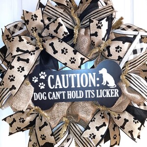 May include: A decorative dog-themed wreath with a black and white sign that reads "CAUTION: DOG CAN'T HOLD ITS LICKER." The wreath features tan and black ribbons with paw prints and bone patterns.