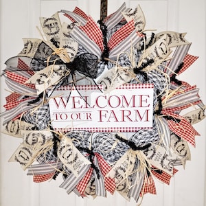 May include: A farmhouse-style wreath with a welcome sign that reads "Welcome to Our Farm". The wreath is decorated with red and white gingham ribbon, black and white buffalo check ribbon, and burlap ribbon with farm-themed prints.