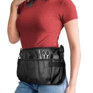 May include: A black stylist's apron with multiple pockets and a variety of tools, including scissors, combs, and clippers. The apron is worn by a person with long black hair wearing a red shirt and blue jeans.