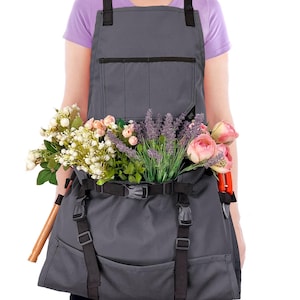 May include: A gray gardening apron with black straps and pockets, filled with an assortment of colorful flowers. The apron has a waist belt and is designed to hold gardening tools. A pair of orange handled shears is visible.