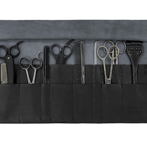 Puede incluir: Rollo de herramientas de barbero de cuero negro con varias tijeras, un peine, una navaja y un cepillo para tinte de pelo. El rollo está abierto, mostrando las herramientas, y tiene un forro de gamuza gris. Una correa negra con hebilla asegura el rollo cuando está cerrado.