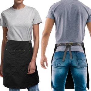 以下が含まれることがあります： 調節可能なストラップと大きなフロントポケットが付いた黒いキャンバスエプロン。エプロンには真鍮のリベットと真鍮のバックルが付いた革のベルトが付いています。