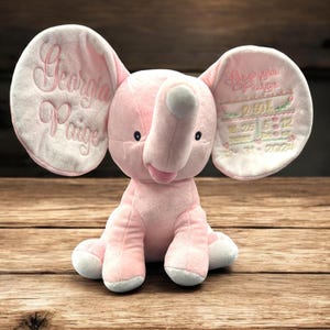 May include: A pink plush elephant with embroidered details. The elephant's ears are embroidered with the name "Georgia Paige" and birth details. The elephant is sitting on a wooden surface.