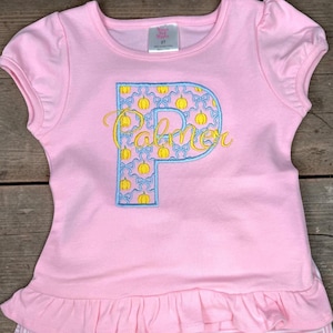 May include: A light pink short-sleeved t-shirt with a ruffled hem. The center features a large letter P appliqued in a light blue fabric with a yellow pumpkin and blue bow pattern. The name Palmer is embroidered in yellow cursive script across the P.
