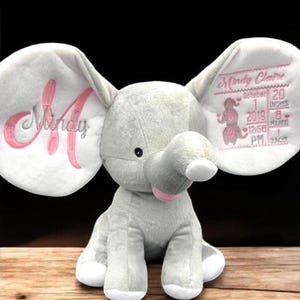May include: A gray plush elephant with large, embroidered ears. The left ear features the name "Mindy" in pink script with a large pink "A". The right ear displays birth details: name, date, time, height (1 inch), weight (8 pounds), and ounces (1 ounce).