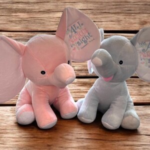 May include: Two plush elephant toys, one pink and one light blue, sit side-by-side on a wooden surface. Each elephant has large ears with embroidered text. The pink elephant's ear reads "All Precious Sight." The blue elephant's ear reads "We All Precious Sight."