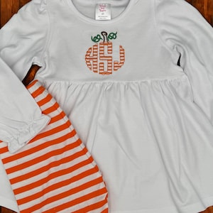 May include: White long-sleeved top with a pumpkin monogram and orange and white striped pants. The pumpkin design is embroidered in orange and green. The top has a gathered waist and the sleeves have a ruffle detail at the wrist. The pants are striped.