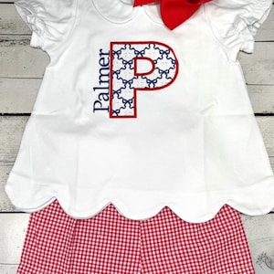 May include: White baby outfit with a red bow. The top has a scalloped hem and a large embroidered letter "P" with a bow pattern. The shorts are red and white gingham with a scalloped edge. The word "Palmer" is embroidered on the top.