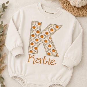 May include: A cream-colored long-sleeve romper featuring a large letter K filled with a pattern of small orange pumpkins and a rope-like design. Below the letter, the name Katie is embroidered in orange cursive script. The romper has ribbed cuffs.