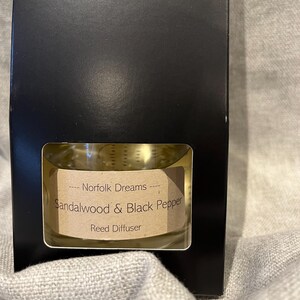 May include: A black box with a clear window showing a glass bottle of reed diffuser. The label on the bottle reads "Norfolk Dreams - Sandalwood & Black Pepper Reed Diffuser".