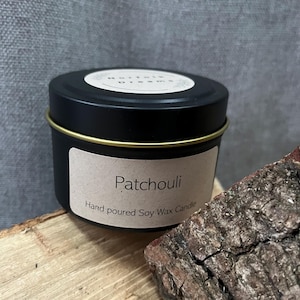 Puede incluir: Un tarro de vela negro con un borde dorado y una etiqueta que dice "Patchouli". La etiqueta también dice "Hand poured Soy Wax Candle". La tapa tiene las palabras "Mornful Dreams". La vela está sobre una superficie de madera.