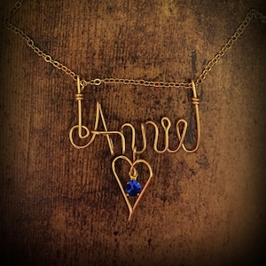 May include: Gold necklace with the name "April" spelled out in wire and a blue gemstone heart pendant.