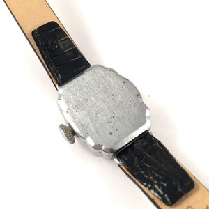 May include: A silver-toned wristwatch with a black leather band. The watch has a square face with rounded edges and a small knob on the side.