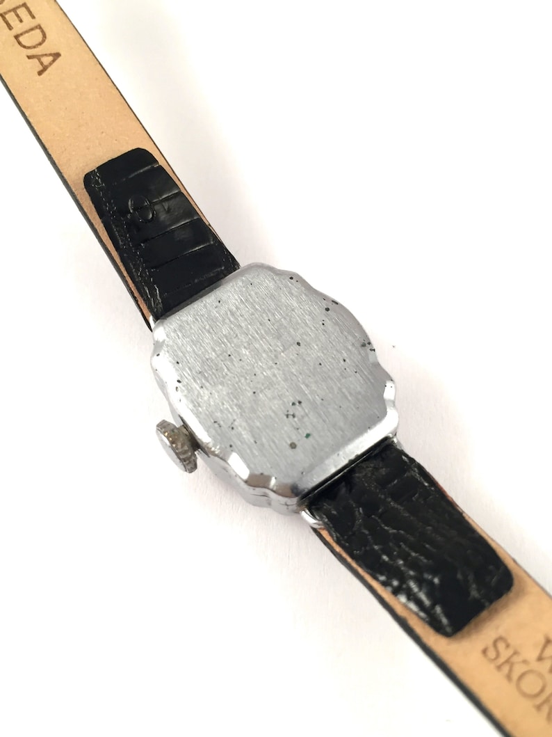 May include: A silver-toned wristwatch with a black leather band. The watch has a square face with rounded edges and a small knob on the side.