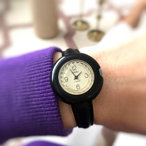 May include: A black wristwatch with a white face and black leather band. The watch face has black Roman numeral hour markers and a black second hand. The watch face also has the text "USSR" and "17 jewels" printed on it.