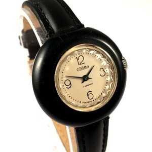 May include: A black leather wristwatch with a round, black face. The face has white numbers and a white dial with the word "Слава" and "17 камней" written on it. The watch has a black leather band.