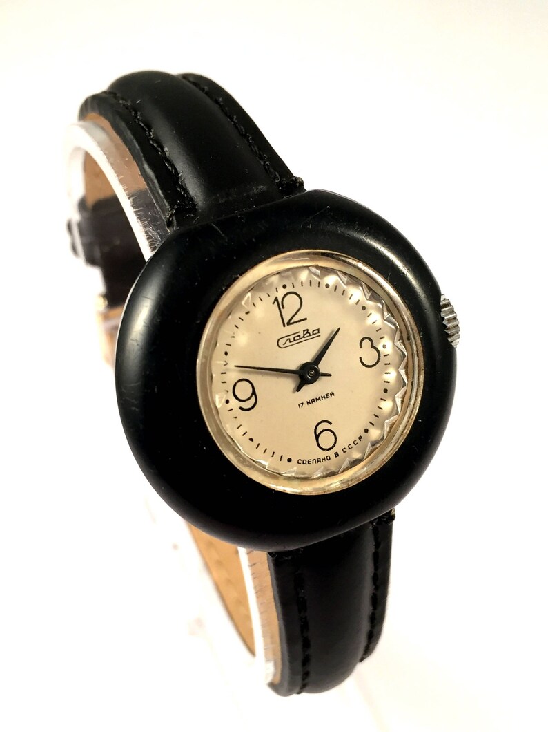 May include: A black leather wristwatch with a round, black face. The face has white numbers and a white dial with the word "Слава" and "17 камней" written on it. The watch has a black leather band.