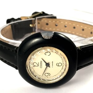 May include: A black wristwatch with a round, black face and a white dial. The watch has a black leather band with white stitching. The dial has gold-colored hour markers and hands. The watch face has the text "СССР" and "Made in USSR" printed on it.