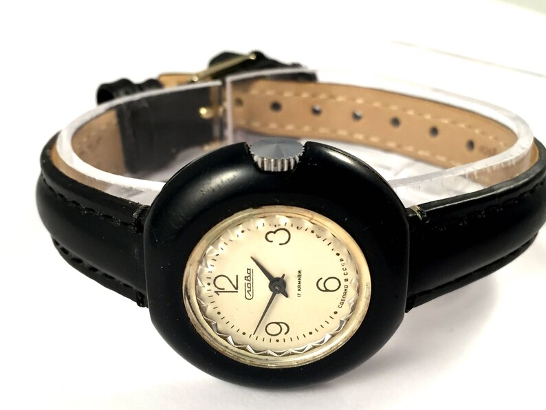 May include: A black wristwatch with a round, black face and a white dial. The watch has a black leather band with white stitching. The dial has gold-colored hour markers and hands. The watch face has the text "СССР" and "Made in USSR" printed on it.