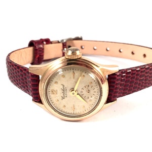 May include: A gold-toned wristwatch with a white face and black hands. The watch has a red leather band with a crocodile pattern. The watch face says "Waltham Sport".