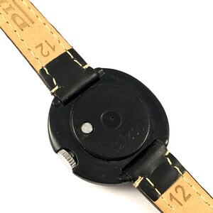 May include: Black round wristwatch with a black leather band. The watch face is covered with a clear plastic cover. The back of the watch has the text "21753" printed on it.